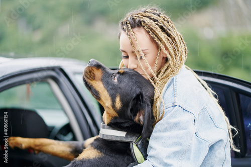 Young woman with blonde braided hair wearing a denim jacket and white jean kissing her dog on a rainy day photo