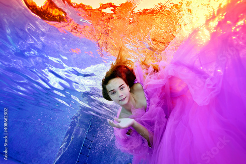 A cute girl in a lush pink dress dives under the water in an outdoor pool. She looks at the camera against the background of the sunset on the surface. Underwater photography. View from the bottom.
