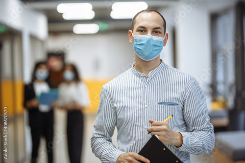 Elegant businessman wearing a medical mask stands in the office corridor with a tablet in his hands. Coronavirus prevention measures, new normal concept. Portrait of a professional worker.