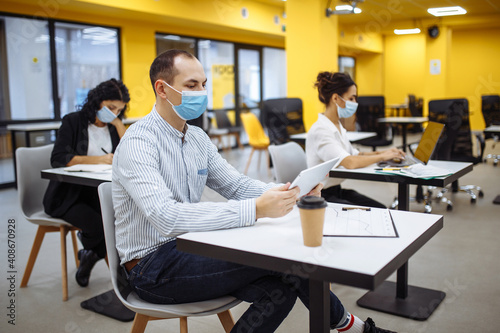 Three colleagues work together at an office keeping social distance weaing medical masks. People sit at the desks working hards to support business. New normal, covid-19 pandemic measures concept.
