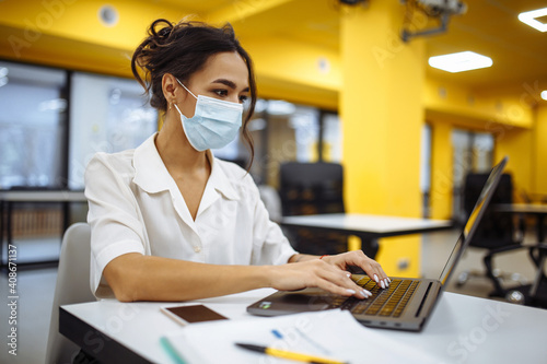 Portrait of a woman working on a laptop at an office desk, wearing medical mask and staying safe during covid-19 pandemic outbreak. Health care and self hygiene concept.