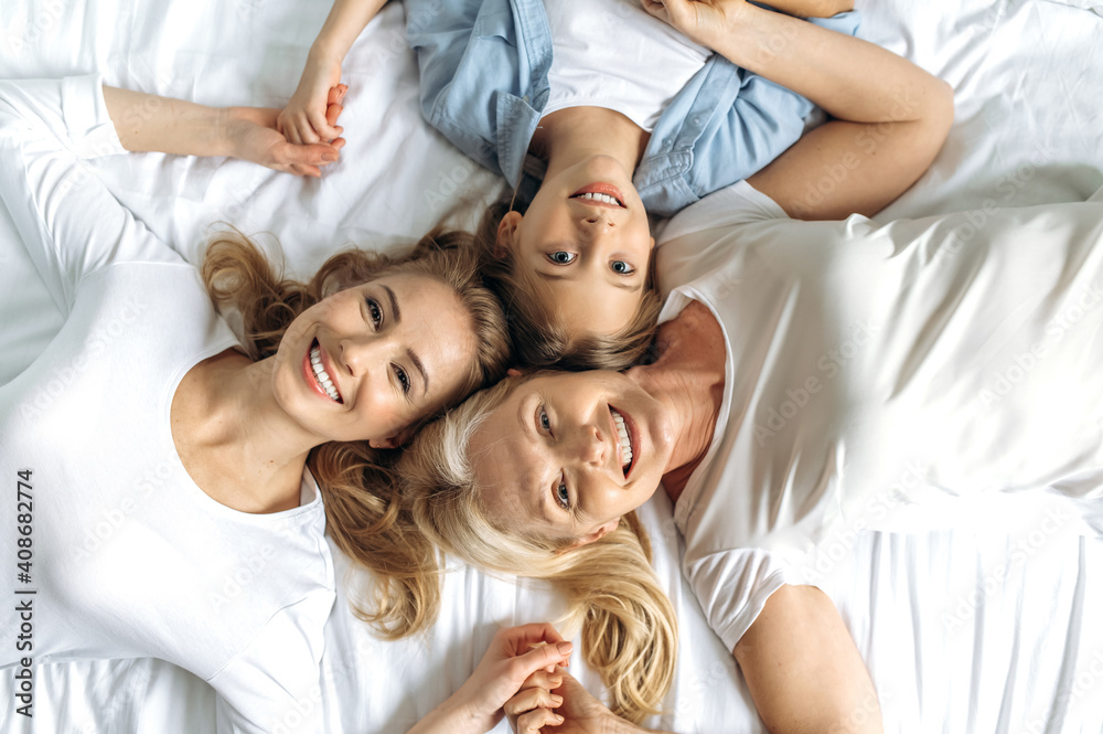 Top view portrait of cheerful happy grandmother daughter and granddaughter lying on the bed holding hands, looking at the camera and smiling. Family relationships and values