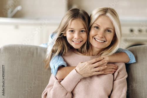 Portrait of a happy caucasian grandmother and granddaughter, or a mom with a daughter, they are hugging sitting on the sofa in the living room, smiling happily and looking at the camera