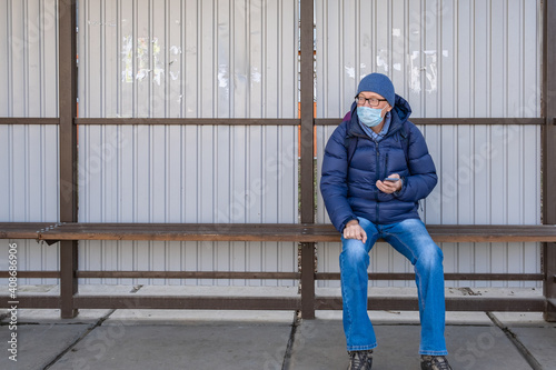 A middle-aged man in a medical mask sits at a public transport stop and waits for a bus. Looks into the distance.