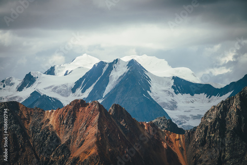 Atmospheric mountains landscape with great snowy top behind colorful brown red orange rocky wall under cloudy sky. Dramatic highland scenery with giant glacier and big vivid brown red orange mountain. © Daniil