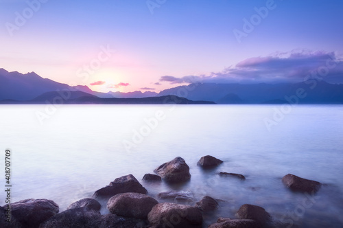 Wonderful sunset at Lake Te Anau - Patience Bay - long exposure with rocks in water and mountain range silhouetted in the background in Fiordland National Park, New Zealand, South Island.
