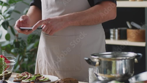 Tilt up shot of male food blogger in apron photographing gourmet toasts on kitchen table with smartphone while preparing content for social media post photo