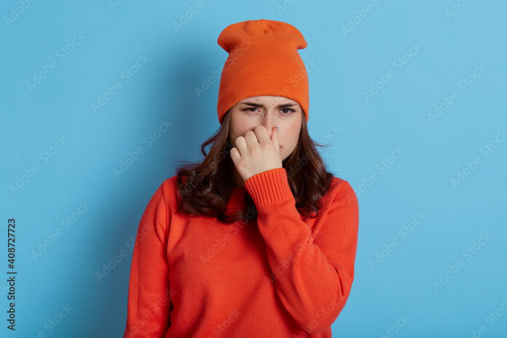 Dissatisfied woman frowns face, lady in cap and casual jumper holds nose because of bad smell, standing isolated over blue background, adorable dark haired girl sees something disgusting. Dissatisfied woman frowns face, lady in cap and casual jumper holds nose because of bad smell, standing isolated over blue background, adorable dark haired girl sees something disgusting.
