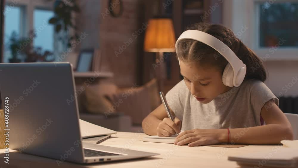 Medium shot of little girl in headphones sitting at desk at home and talking on video call with teacher while studying online Medium shot of little girl in headphones sitting at desk at home and talking on video call with teacher while studying online