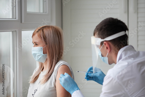 A doctor in a protective suit holds a syringe in rubber gloves and vaccinates a woman patient in a medical mask. Covid-19 or coronavirus vaccine.