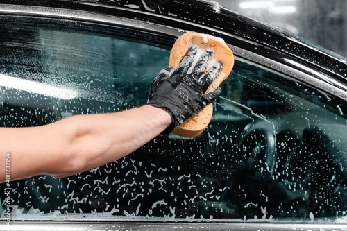 Close up of car wash worker wearing protective gloves and washing car window with soapy sponge. Cleaning services.
