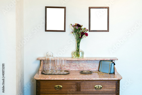 Vintage commode with marble counter top, flowers, hand bag and empty frames on the wall. Luxury home interior