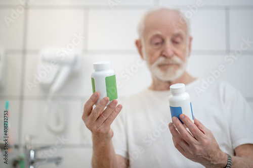 Gray-haired man in white tshirt holding bottles with pills and looking thoughtful