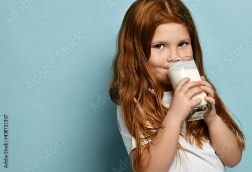 Obraz na płótnie Portrait of happy red haired kid girl drinking milk or yogurt and looking at camera over blue background with free copy space. Health and diet concept. Healthy drink breakfast and clean food concept w salonie