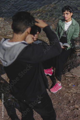 portrait of a female model sitting on a rock near lake wearing green jacket , photographer in black hoodie clicking photos 