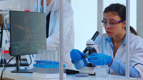 Scientist making research using microscope while colleagues working on background in modern equipped laboratory. Multiethnic stuff analysing vaccine evolution using high tech for researching treatment