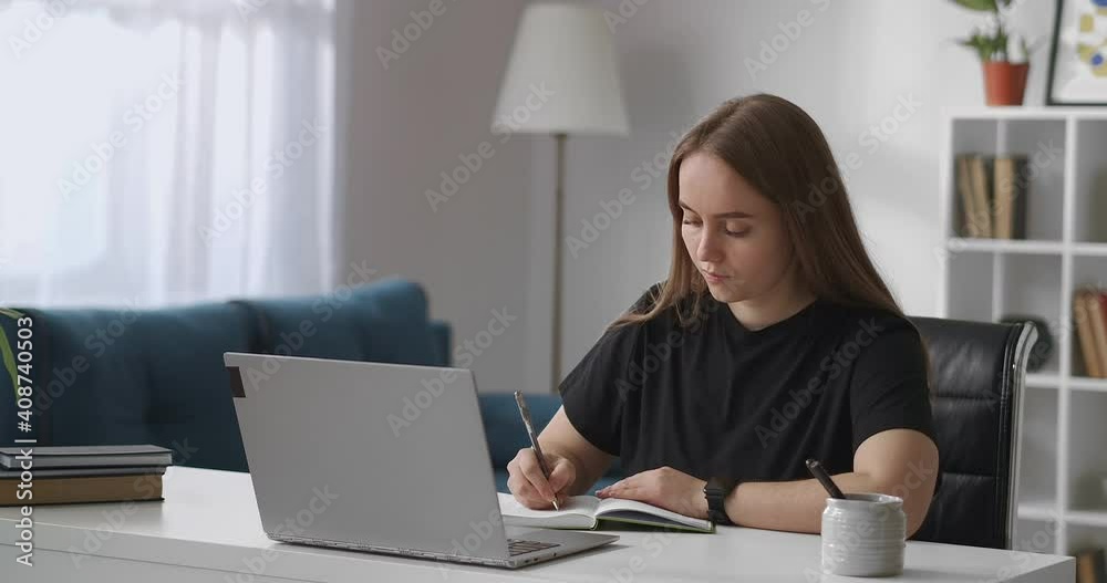 self-education by internet, woman is viewing learning video on display of laptop and writing notes in notebook, sitting at home self-education by internet, woman is viewing learning video on display of laptop and writing notes in notebook, sitting at home