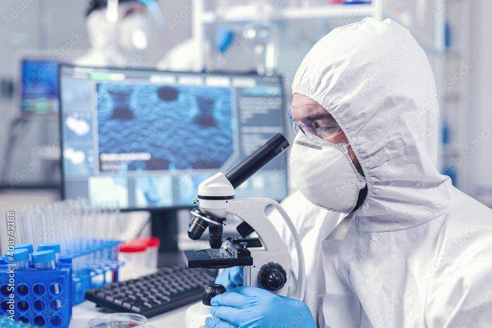 Concentrated scientist in ppe equipment looking into the microscope in laboratory. Scientist in protective suit sitting at workplace using modern medical technology during global epidemic. Concentrated scientist in ppe equipment looking into the microscope in laboratory. Scientist in protective suit sitting at workplace using modern medical technology during global epidemic.