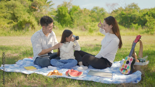 Happy family asian and little girl playing the film camera and have enjoyed ourselves together during picnicking on a picnic cloth in the green garden. Family enjoying sunny fall day in nature. © Kiattisak