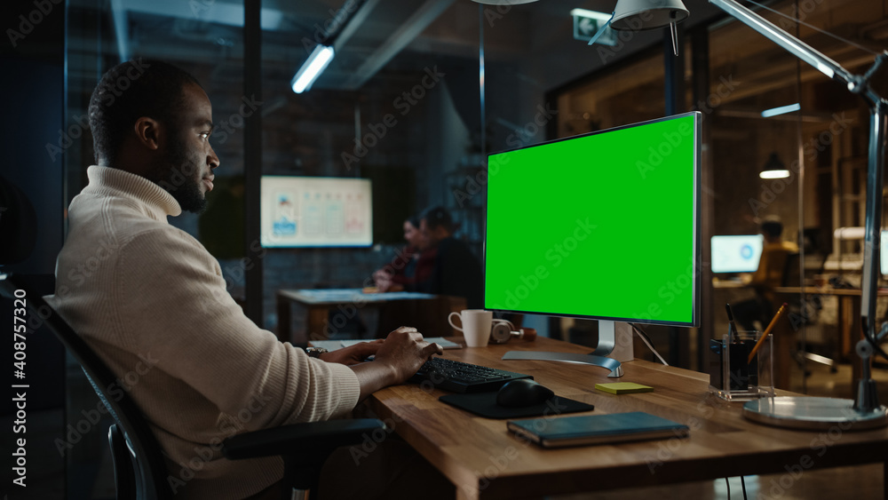 Handsome Black African American Project Manager is Making a Video Call on Desktop Computer with Green Screen Mock Up Display in a Busy Creative Office. Male Specialist is Wearing Turtle Neck Sweater.