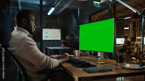 Handsome Black African American Project Manager is Making a Video Call on Desktop Computer with Green Screen Mock Up Display in a Busy Creative Office. Male Specialist is Wearing Turtle Neck Sweater.