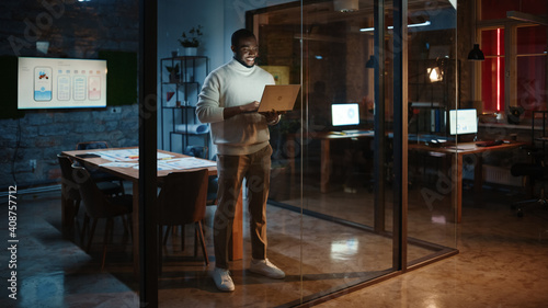 Handsome Black African American Male is Making a Video Call while Standing in Meeting Room Behind Glass Walls with a Laptop Computer in an Creative Agency. Project Manager Wearing White Jumper. © Gorodenkoff