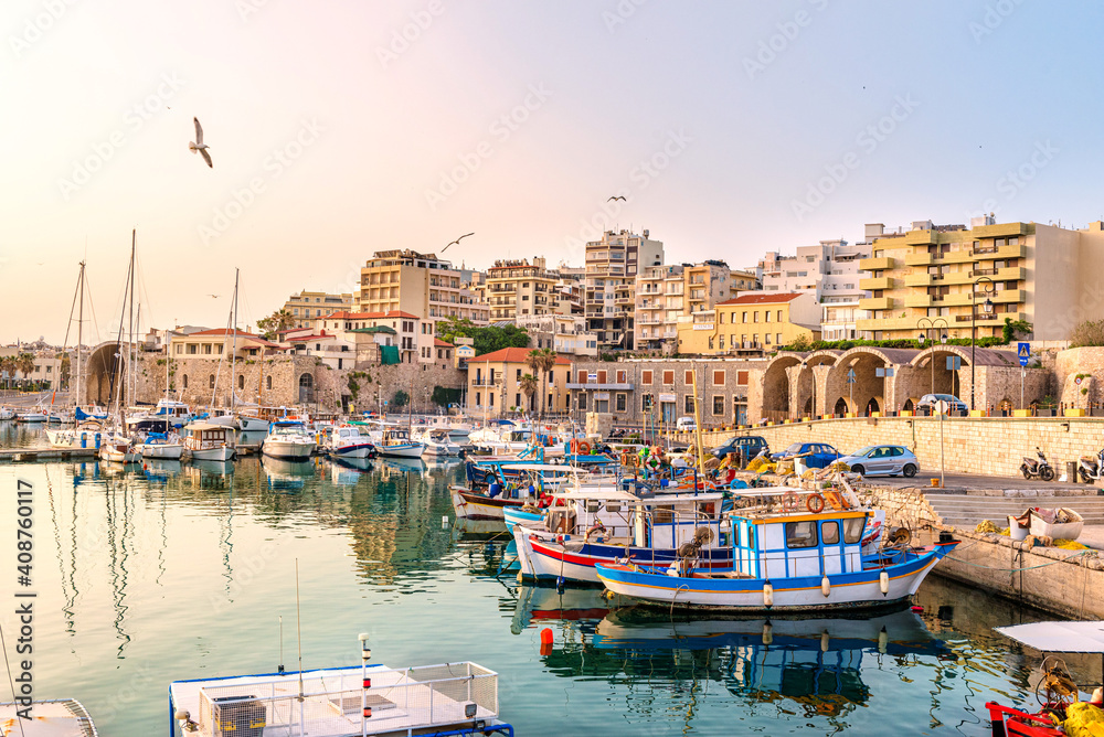 View to Heraklion in the early morning during sun rise, the old port with traditional fishing boats and town with its antient buildings at the background. Selective focus