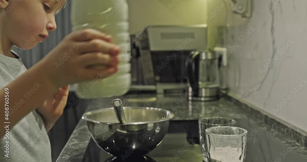 Сhild takes a bottle of milk and pours it into an iron bowl. 8-year-old white boy cooks in the kitchen. Mealtime. Concept of childhood Сhild takes a bottle of milk and pours it into an iron bowl. 8-year-old white boy cooks in the kitchen. Mealtime. Concept of childhood