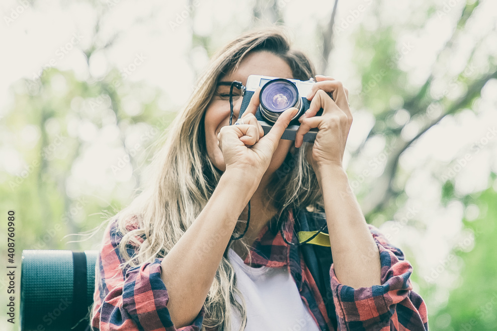 Cheerful blonde woman taking photo of nature with camera and smiling. Caucasian long-haired female tourist trekking in woods. Blurred background. Tourism, adventure and summer vacation concept