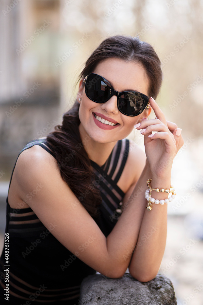Cheerful elegantly beautiful brunette woman, in elegant black dress and sunglasses, posing outside, touching her hands together on the balcony. Cheerful elegantly beautiful brunette woman, in elegant black dress and sunglasses, posing outside, touching her hands together on the balcony.