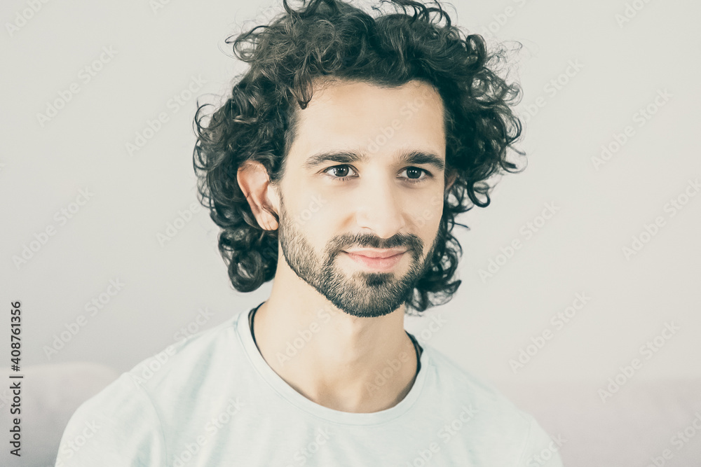 Happy handsome curly haired young man wearing casual t-shirt, sitting on couch at home, looking away and smiling. Male portrait concept