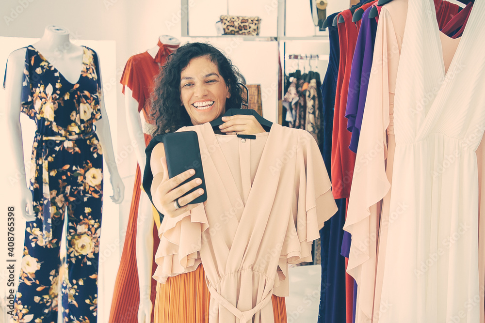 Joyful woman shopping in clothes store and consulting friend on cellphone, showing dress on hanger at frontal camera. Boutique customer or communication concept