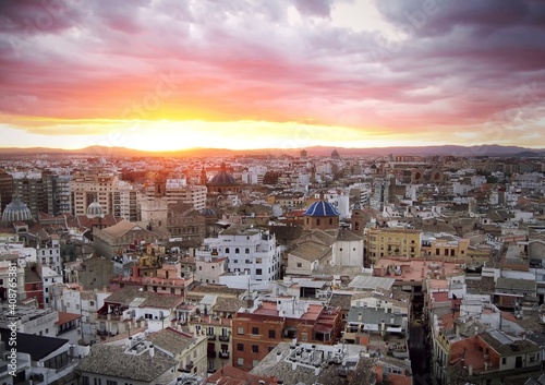 Cityscape of historic Valencia old town against a glowing sunset sky with orange-pink thunderstorm clouds over the rooftops. Narrow streets in the foreground.