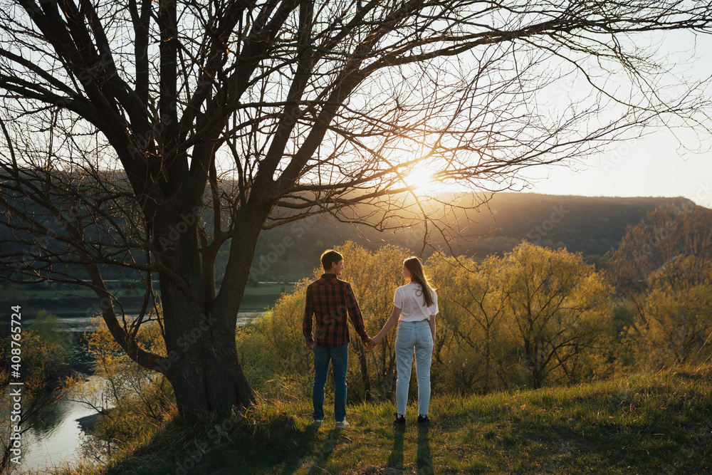 Couple of man and woman holding hands, looking each other and standing together under tree with sunset sky background. Couple of man and woman holding hands, looking each other and standing together under tree with sunset sky background.