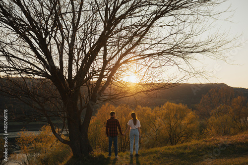 Young couple holding hands while standing under the big tree on hill during sunset in spring time.