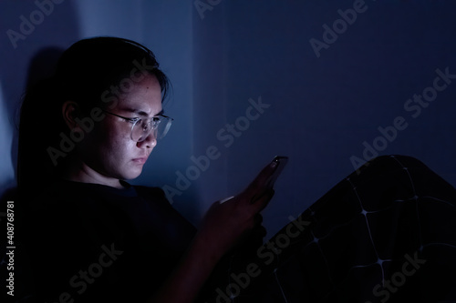 An Asian woman wearing glasses is using a smartphone with a bright screen in the dark.