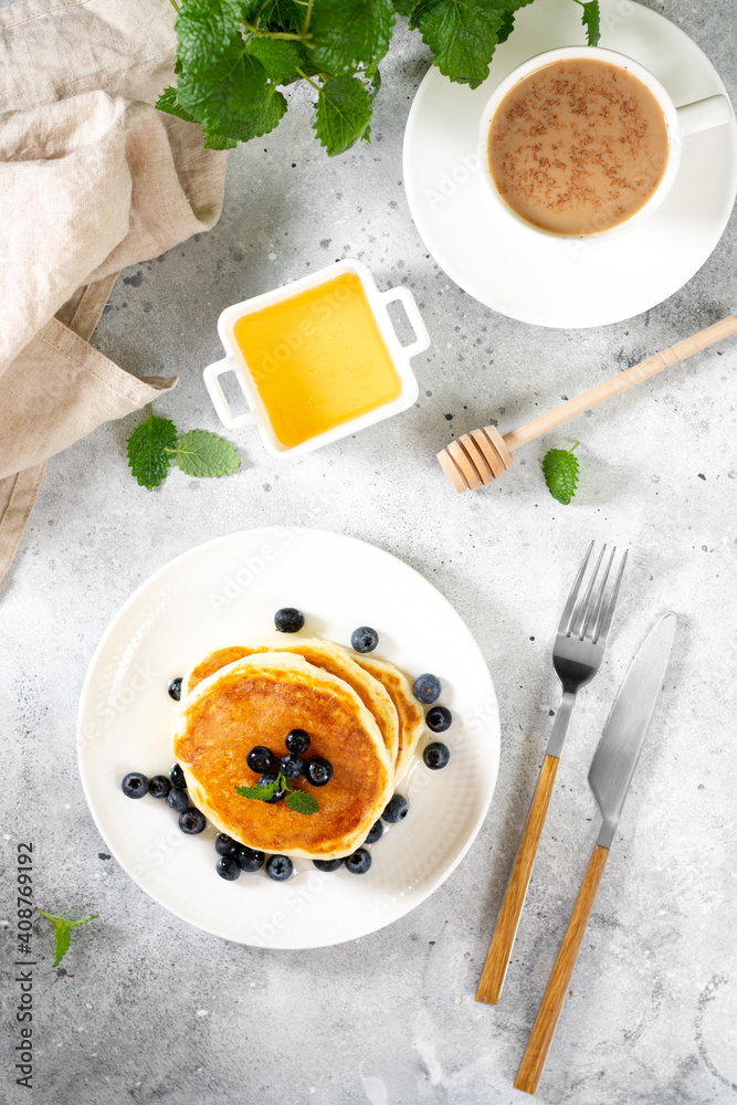 Pancakes in a white plate on the light gray kitchen table. Homemade delicious pancakes with honey and blueberries. Delicious sweet and hearty breakfast Pancakes in a white plate on the light gray kitchen table. Homemade delicious pancakes with honey and blueberries. Delicious sweet and hearty breakfast