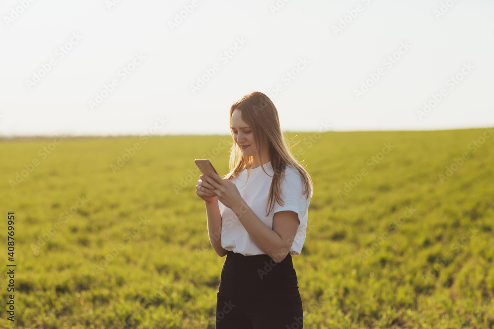 Young girl in white t-shirt laughing and typing message on smartphone in the green field during spring or summer.