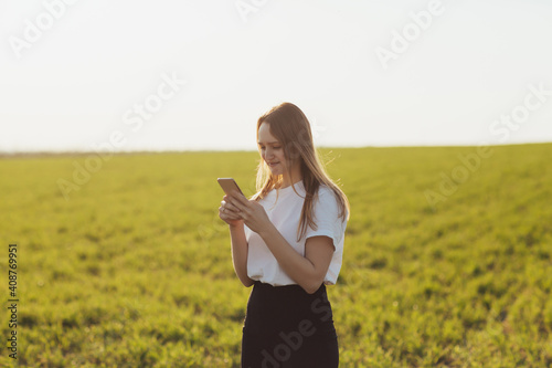 Young girl in white t-shirt laughing and typing message on smartphone in the green field during spring or summer.