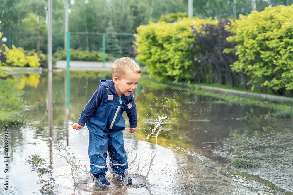 Little cute playful caucasian blond toddler boy enjoy have fun playing jumping in dirty puddle wearing blue waterproof pants and rubber rainboots at home yard street outdoor. Happy childhood concept Little cute playful caucasian blond toddler boy enjoy have fun playing jumping in dirty puddle wearing blue waterproof pants and rubber rainboots at home yard street outdoor. Happy childhood concept