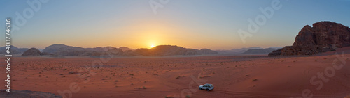 Panoramic view of the sunset in the desert of Wadi Rum, Jordan. The sun sets behind the mountains as a jeep takes in the view.