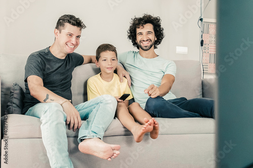 Happy gay fathers and son watching funny TV show at home, sitting on couch in living room, smiling and laughing. Family and home entertainment concept