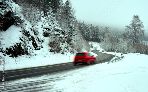 Winding cleared gray asphalt road in Harz mountains with fresh snow on the sides and a red car, intentional motion blur