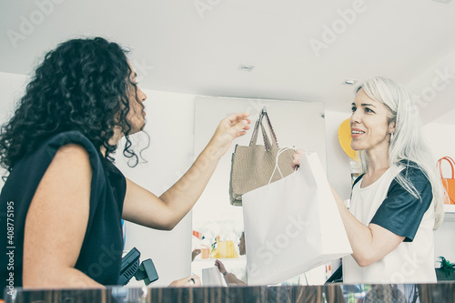 Black haired clothes shop cashier giving paper bag to customer over desk with cash register. Side view. Shopping or consumerism concept