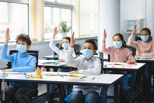Schoolchildren raising hands at classroom, wearing medical masks