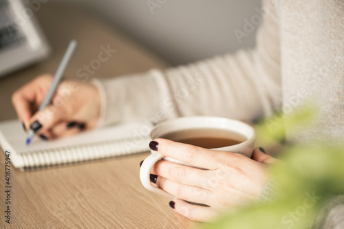 Woman in beige sweater with a tea writing in notebook. 