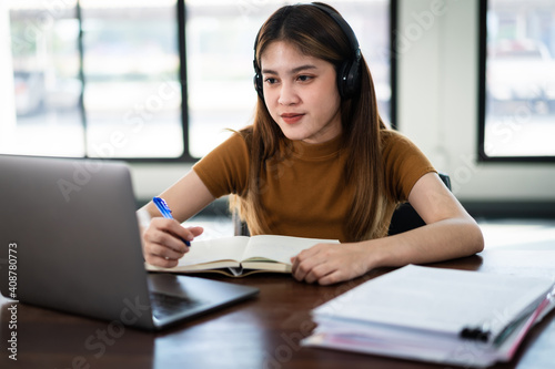 Young  Asian girl student wears wireless headphones concentrates write on the notebook to study language online watch and listen to the lecturer, webinar via video call e-learning at home
