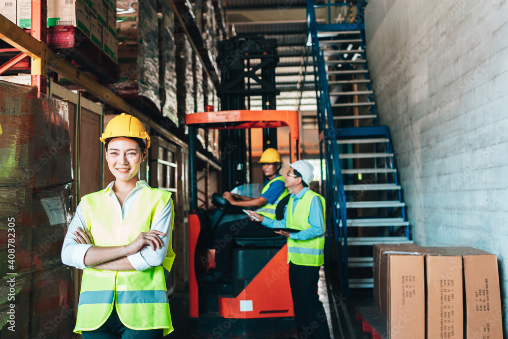Obraz premium Portrait of asian woman. warehouse Smiling worker standing with arms crossed in large warehouse with goods. in background driver at Warehouse forklift loader works with goods.