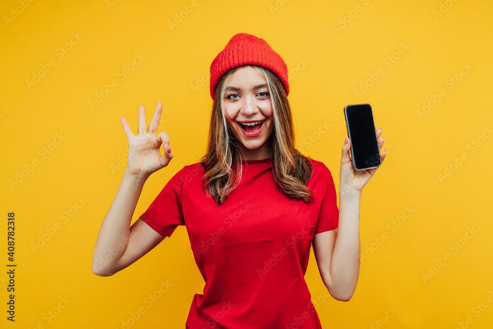 Close up photo of a pretty girl on a yellow background in the studio, she holds a phone with a black screen in her hand and the other hand shows okay. She is dressed in bright red clothes Close up photo of a pretty girl on a yellow background in the studio, she holds a phone with a black screen in her hand and the other hand shows okay. She is dressed in bright red clothes