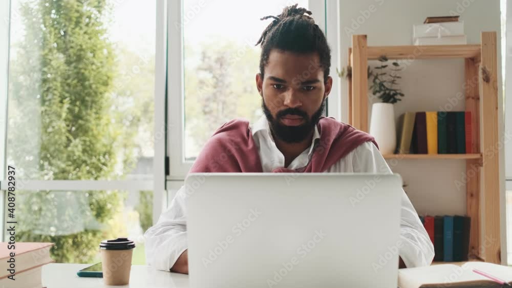 A serious african american man is working with his computer sitting at the table in the office